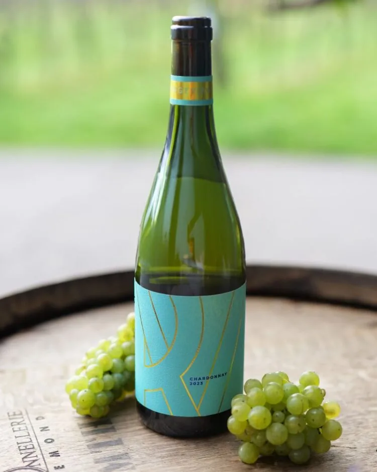 A bottle of Ridgeview's still Chardonnay with a bright blue label, on an oak barrel with Chardonnay grapes and the vineyard in the background.