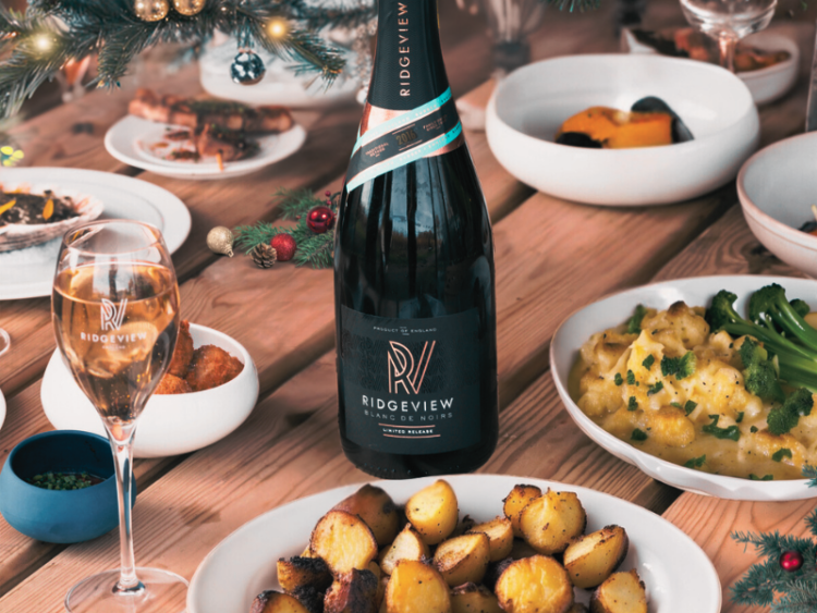 A bottle of Blanc de Noirs in the middle of a table surrounded by plates of Christmas roast dinner and christmas decorations, with a glass of sparkling wine on the table.