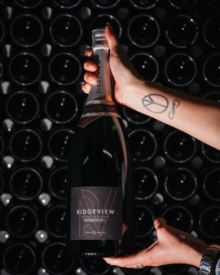 Someone holding up a magnum in the Ridgeview cellar with a tattoo on their arm in front of bottle resting on their side